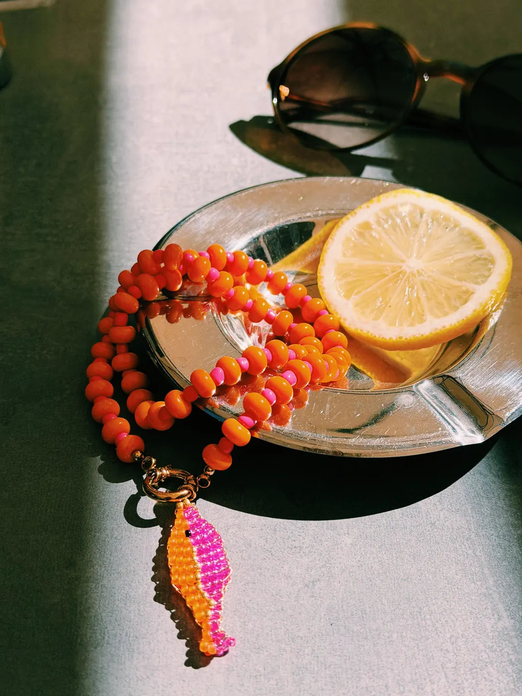 Collier Sorbet mandarine et Grigri poisson posés au soleil, avec une tranche de citron